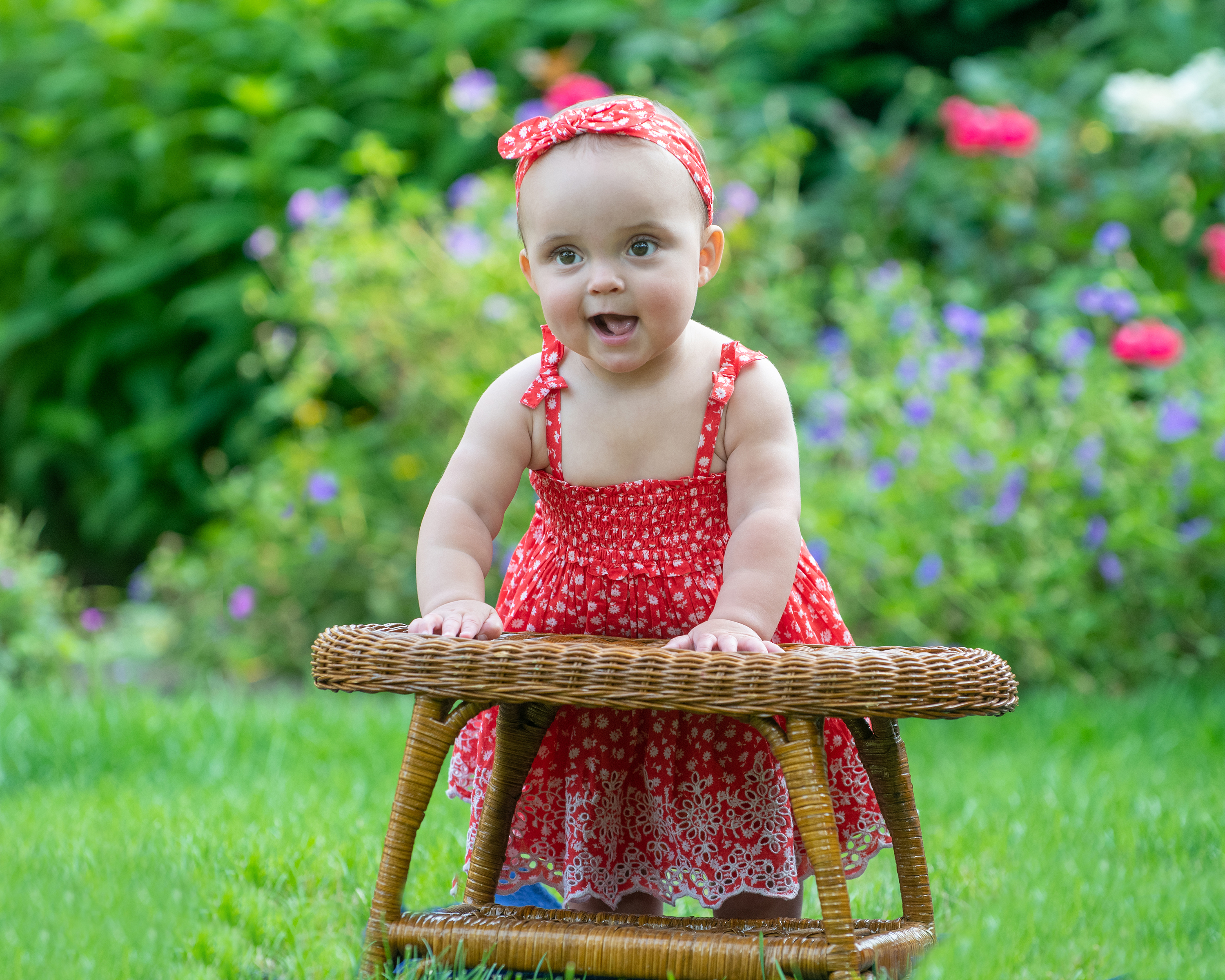 Smiling baby girl in red floral dress on wicker stool in garden.