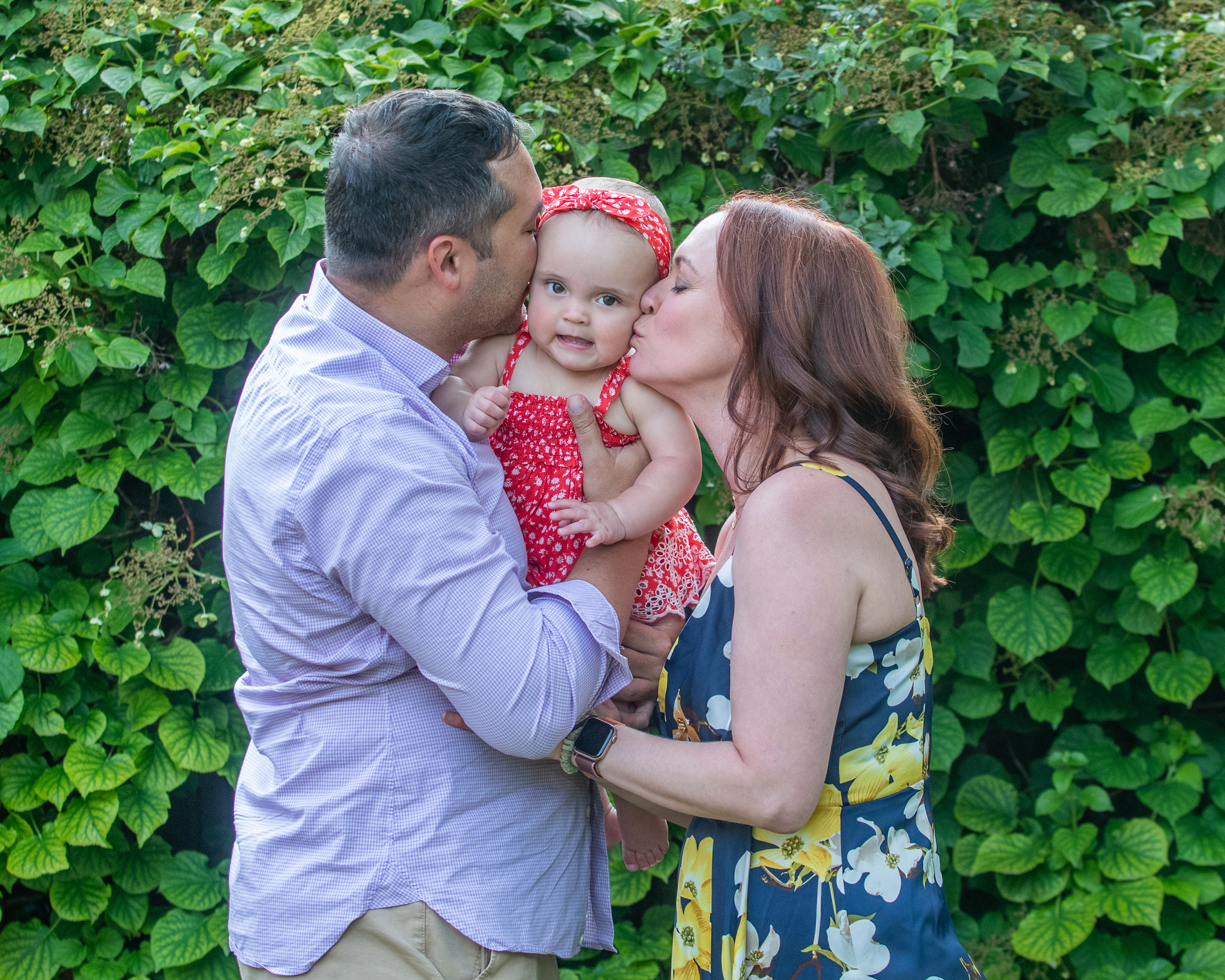 Parents kiss their baby girl on the cheeks while holding her in front of a leafy green backdrop.