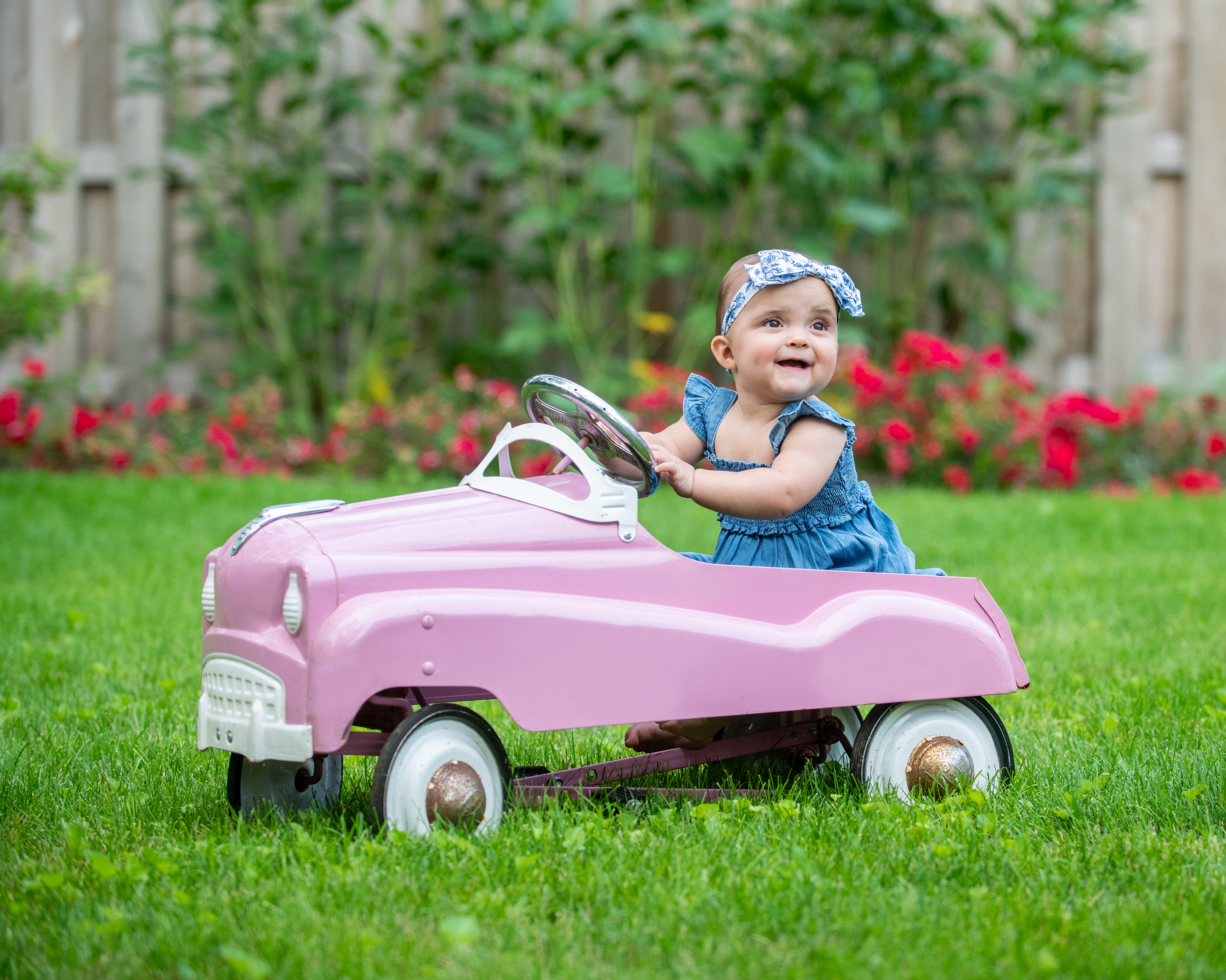 Smiling baby in a blue dress sits in a pink toy pedal car on a grassy lawn with flowers in the background.