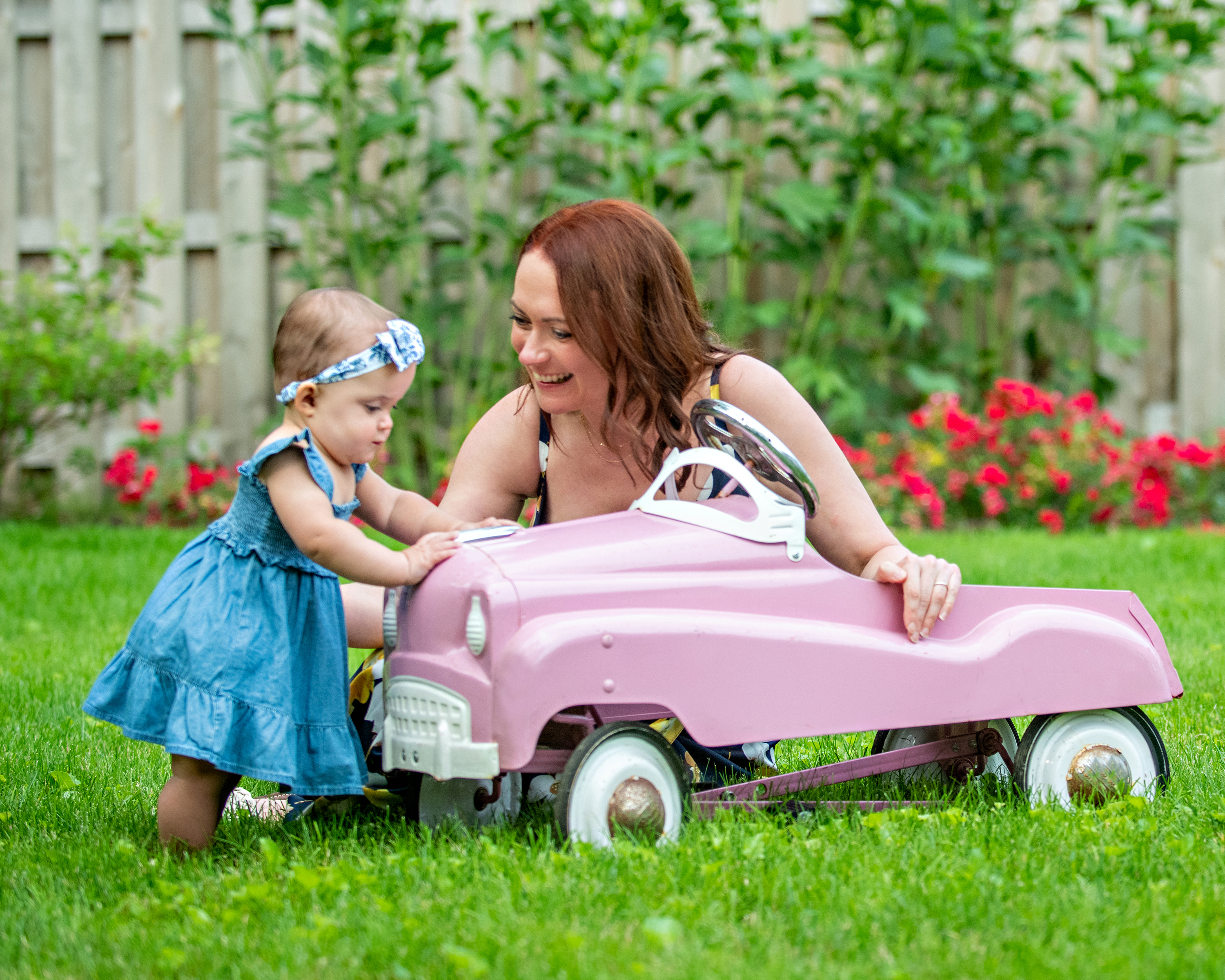 Smiling red-haired mother helping her baby girl in a blue dress with a pink toy car in a grassy backyard.