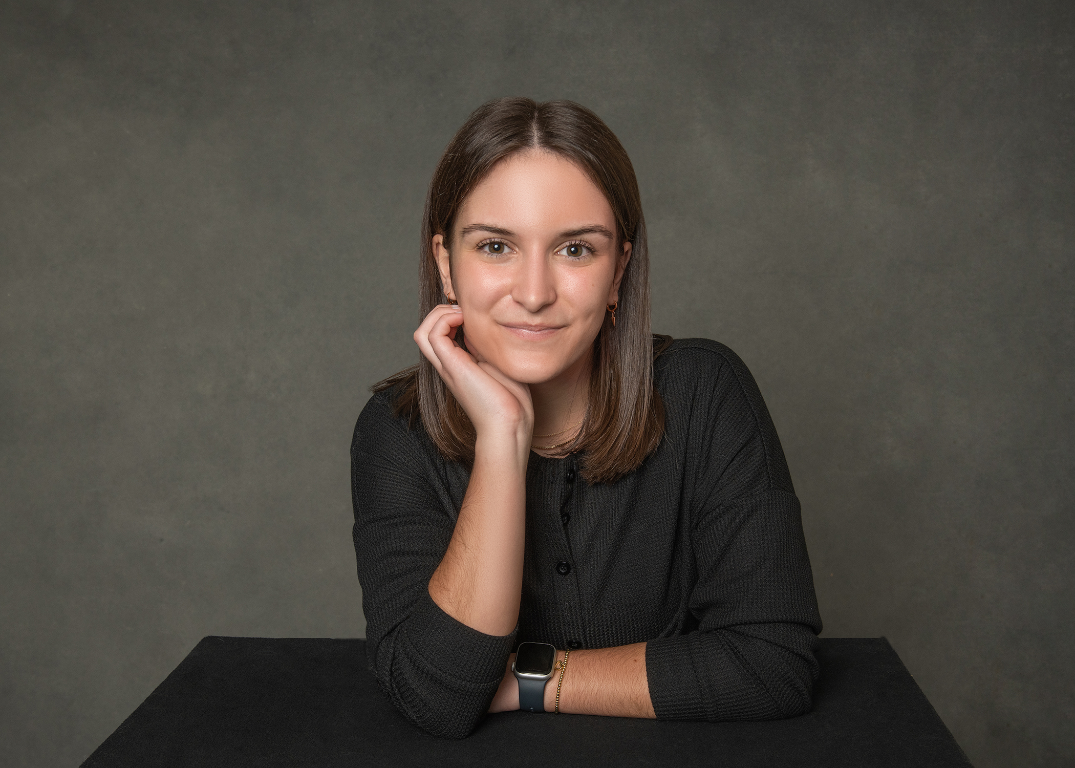Young woman with brown hair in black cardigan rests chin on hand at dark table, smiling warmly at camera against gray studio backdrop.
