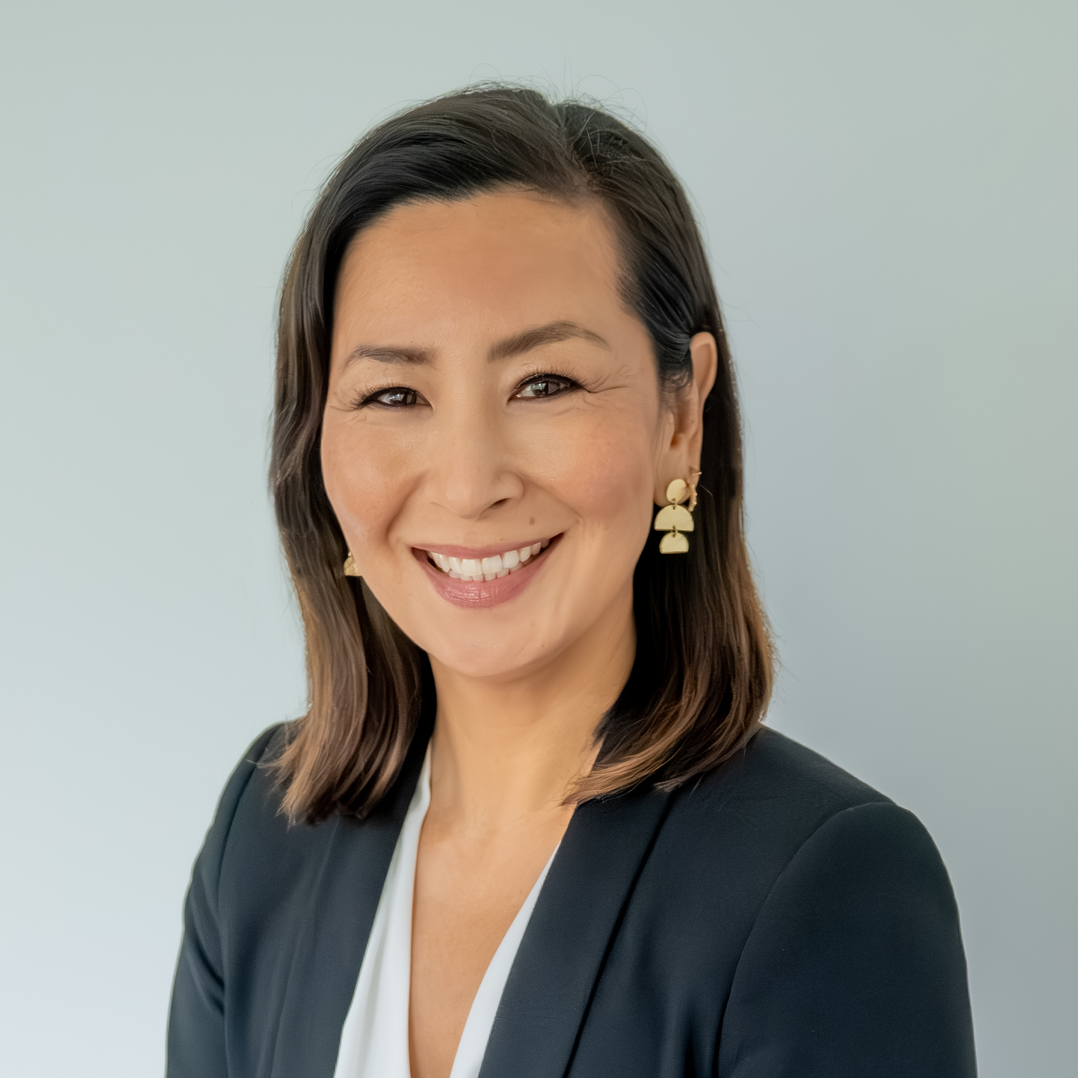Professional woman smiling in a dark blazer and white top with gold earrings against a light gray studio background.