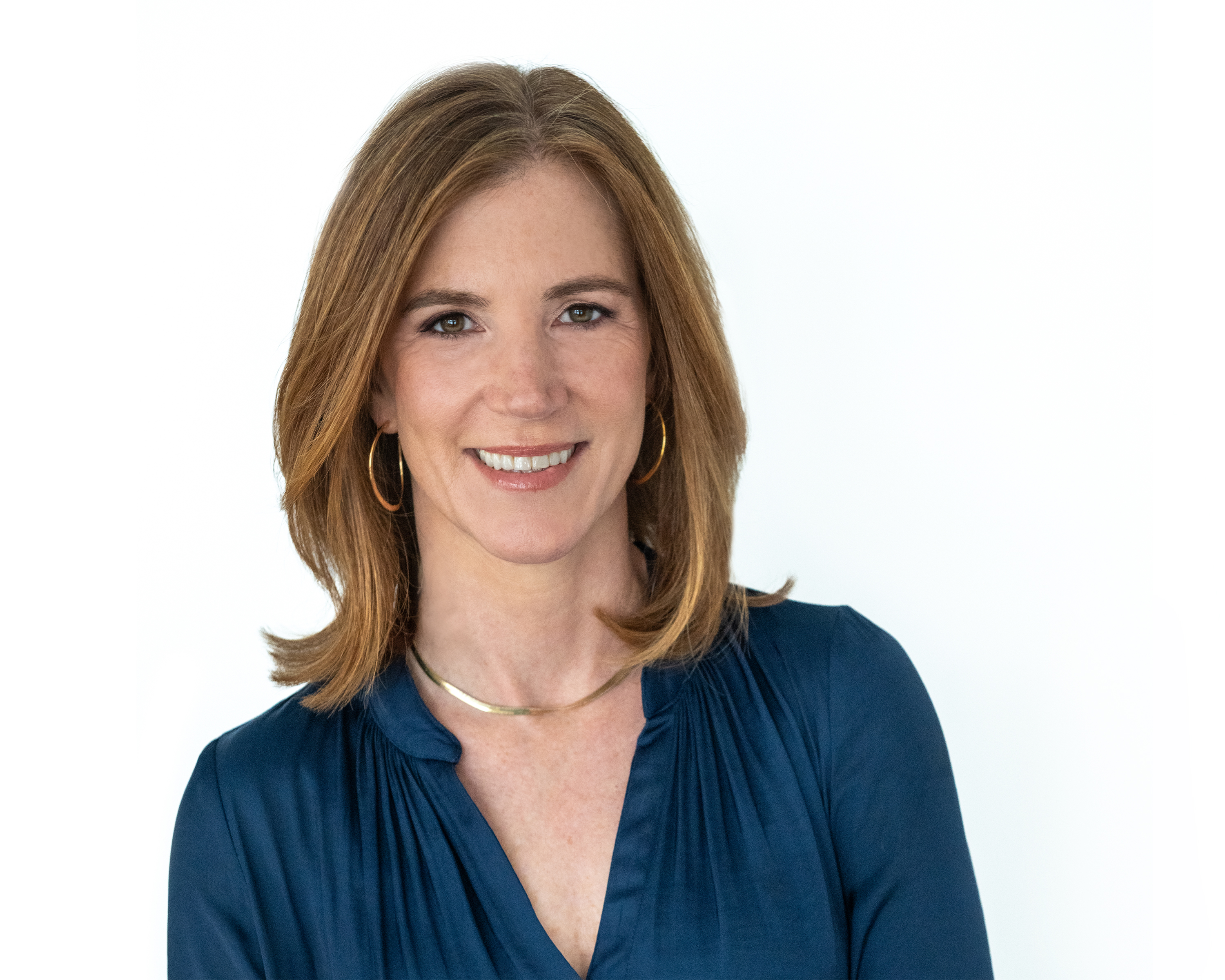 Woman with auburn hair smiling confidently in a navy blouse and gold jewelry against a white background, professional headshot.