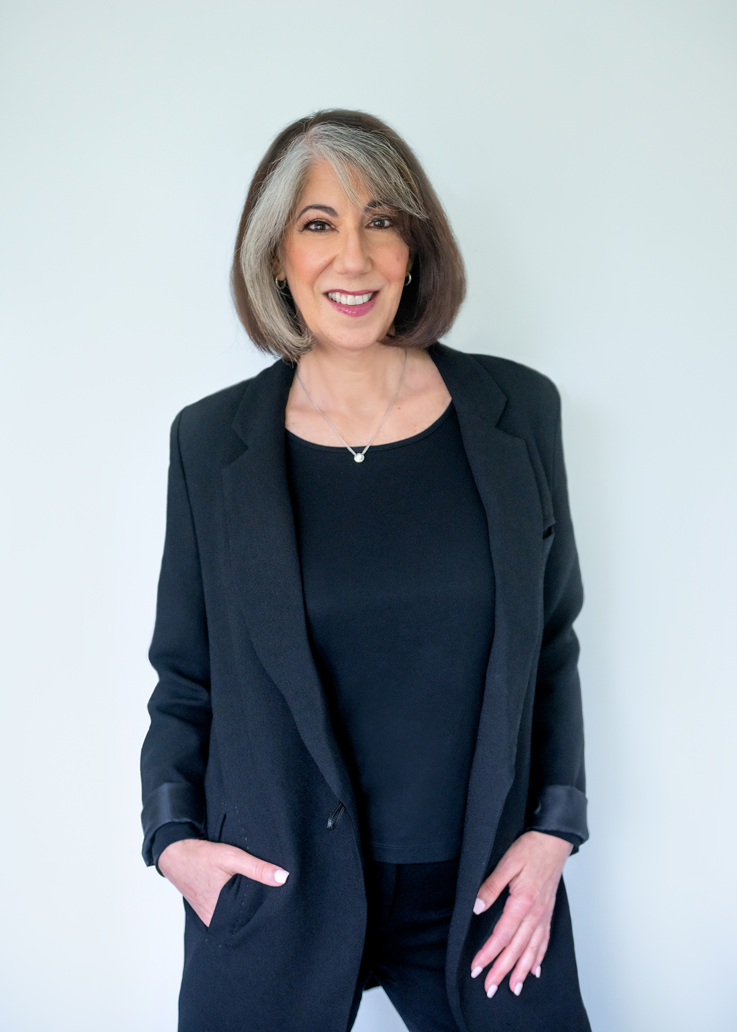 Professional woman in black blazer and top smiling confidently against a light gray studio background.